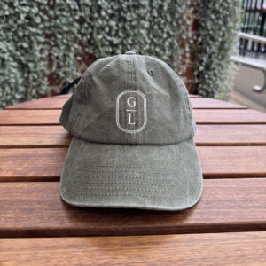 Olive green dad cap with white embroidered GL monogram logo, sitting on a timber table outdoors.